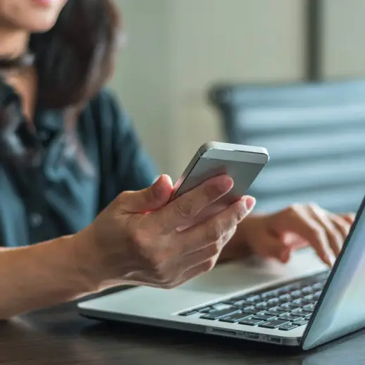 female holding mobile phone while looking at laptop