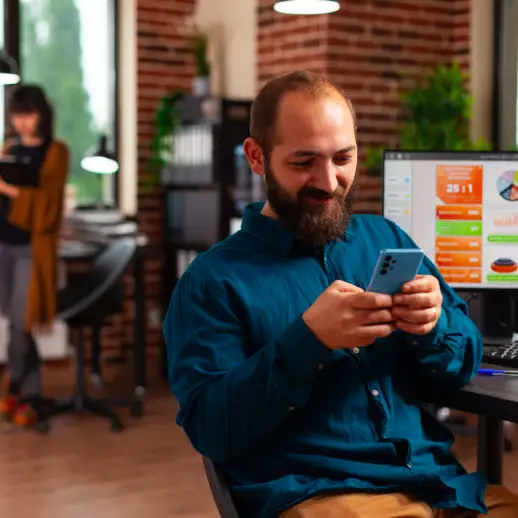 male looking at mobile phone at desk with office background and another employee in background